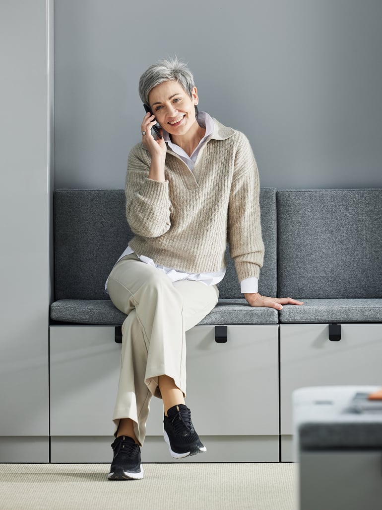Moteris sėdi ant biuro spintelės ir kalbasi telefonu
