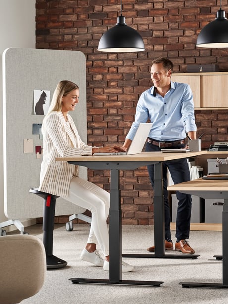 To kolleger taler sammen ved et hæve sænkebord i et kontor med en rustik murstensvæg