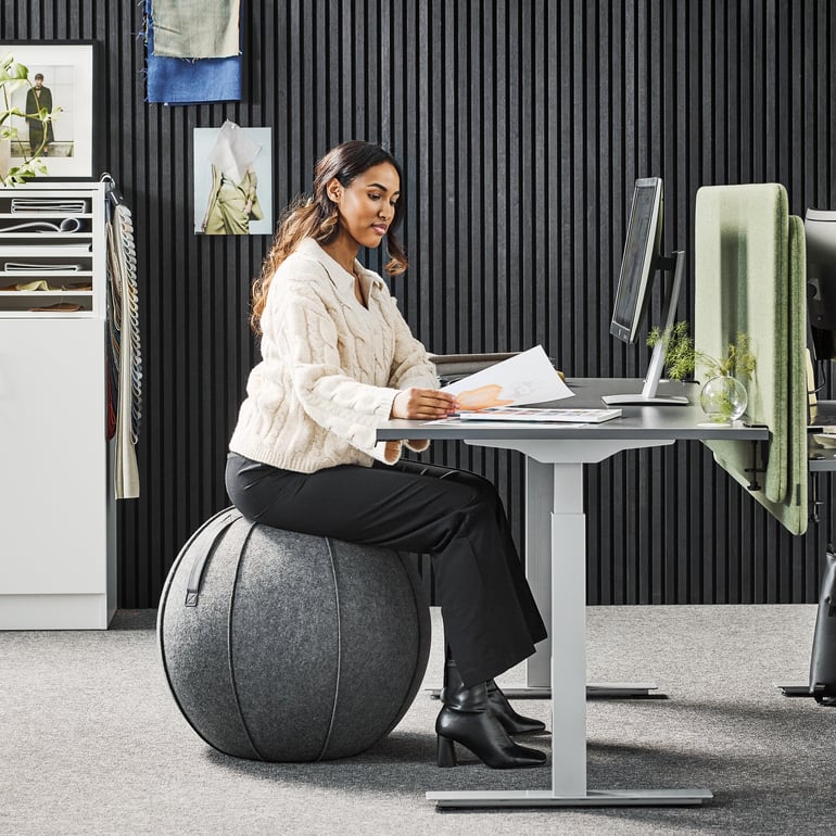 Eine Frau sitzt an einem Sitzball im Büro an ihrem höhenverstellbaren Schreibtisch