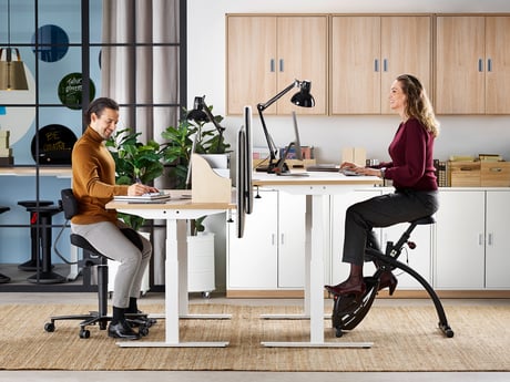 Dvije osobe sjede za elektropodiznim stolom i koriste stolicu sedlo i stolni bicikl za vježbanje