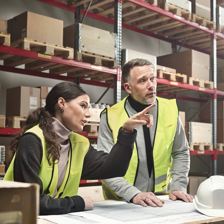 Two people in a warehouse wearing hi-vis jackets and looking at building plans